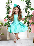 A girl sitting on a swing wearing a mint green paisley pleated dress with short sleeves and a pleated skirt. The dress features a back zipper and is made of a fabric that appears to be a blend of cotton and polyester.