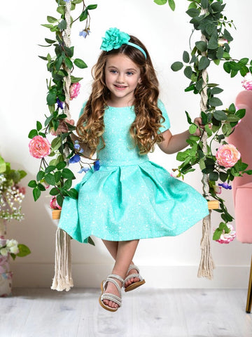 A girl sitting on a swing wearing a mint green paisley pleated dress with short sleeves and a pleated skirt. The dress features a back zipper and is made of a fabric that appears to be a blend of cotton and polyester.