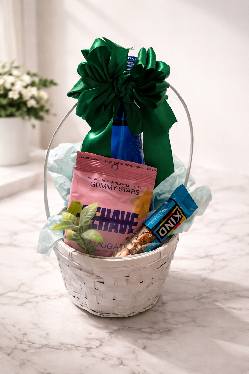 White woven gift basket with green satin bow featuring Saratoga water, Behave zero sugar gummy candy, and a KIND bar with decorative greenery.